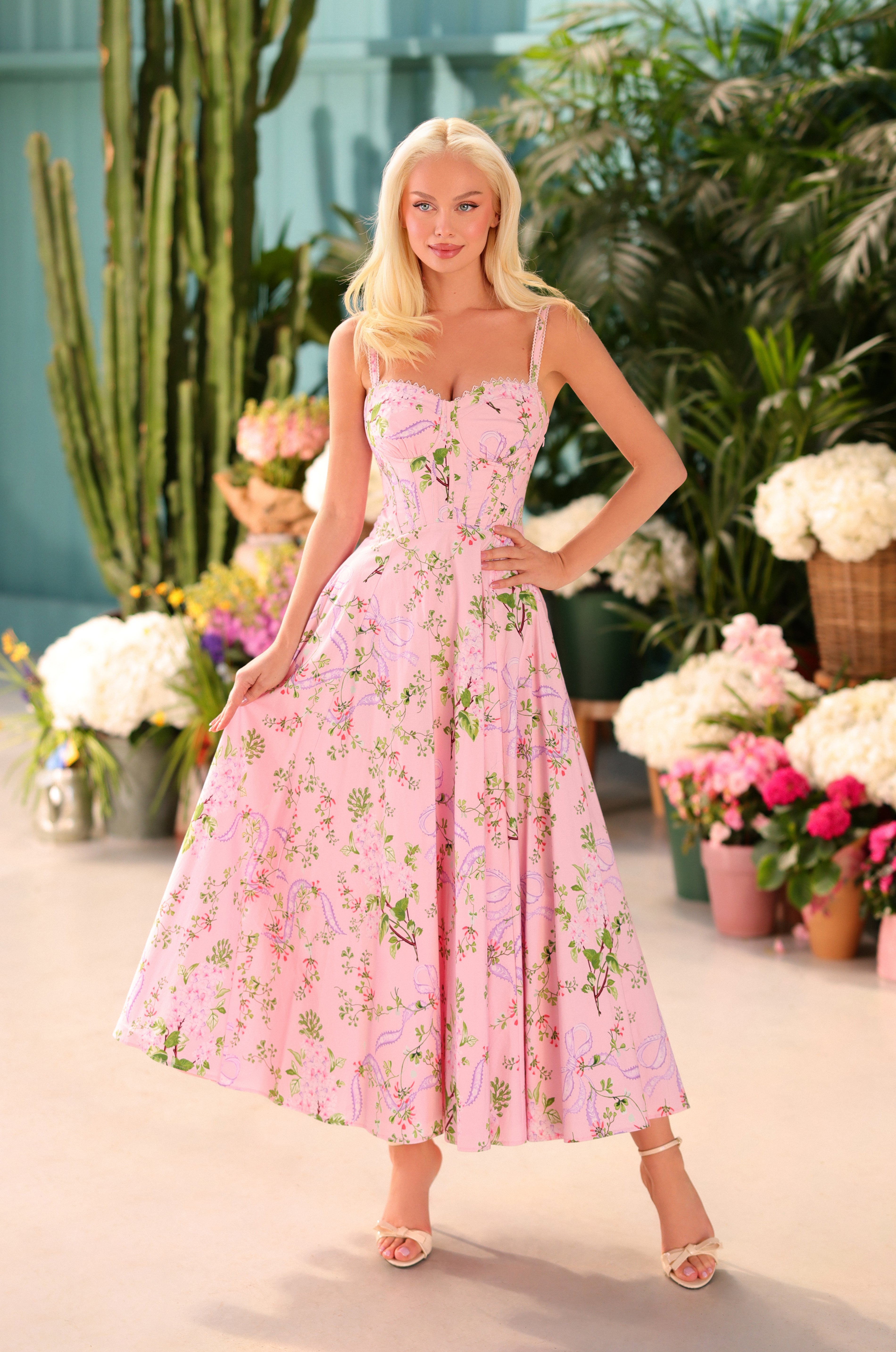 A woman stands indoors among green plants and colorful flowers, wearing the Rosella blush pink floral & bow print casual dress.